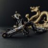 Automobilių pakabukas „Rytietiški pažadai Alba“ – kryžiaus formos pakabukas iš arti, rankų darbo juodi ornamentai ir aukso detalės, fone rytietiškas drakono skulptūros motyvas